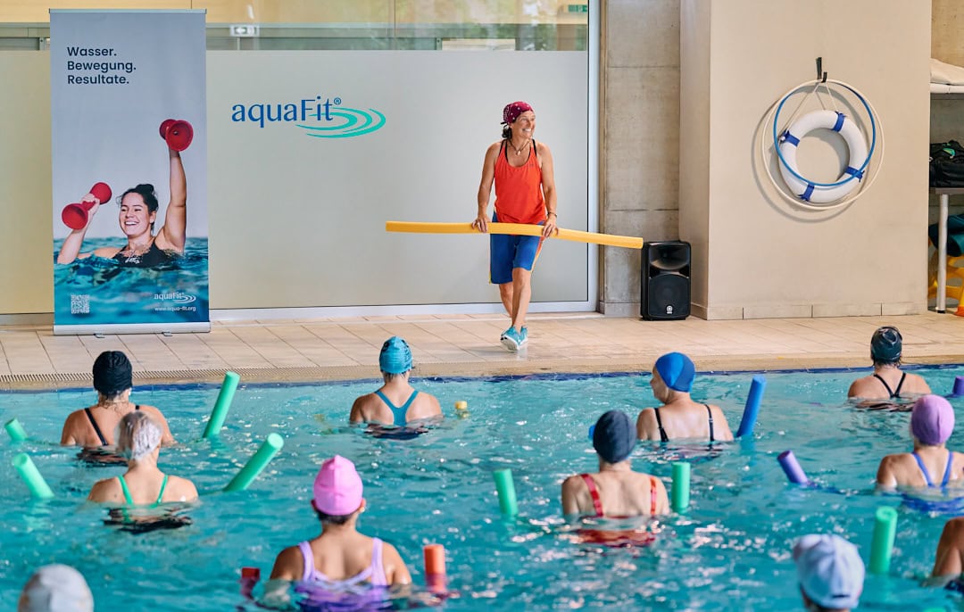 Aqua Fit Wassertraining mit Trainerin im Schwimmbad, Teilnehmer mit Poolnudeln, Wassersport für Fitness und Gesundheit.
